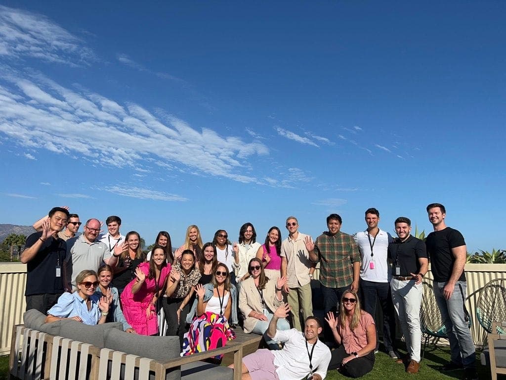 About twenty-five team members waving on a rooftop terrace under a blue sky.
