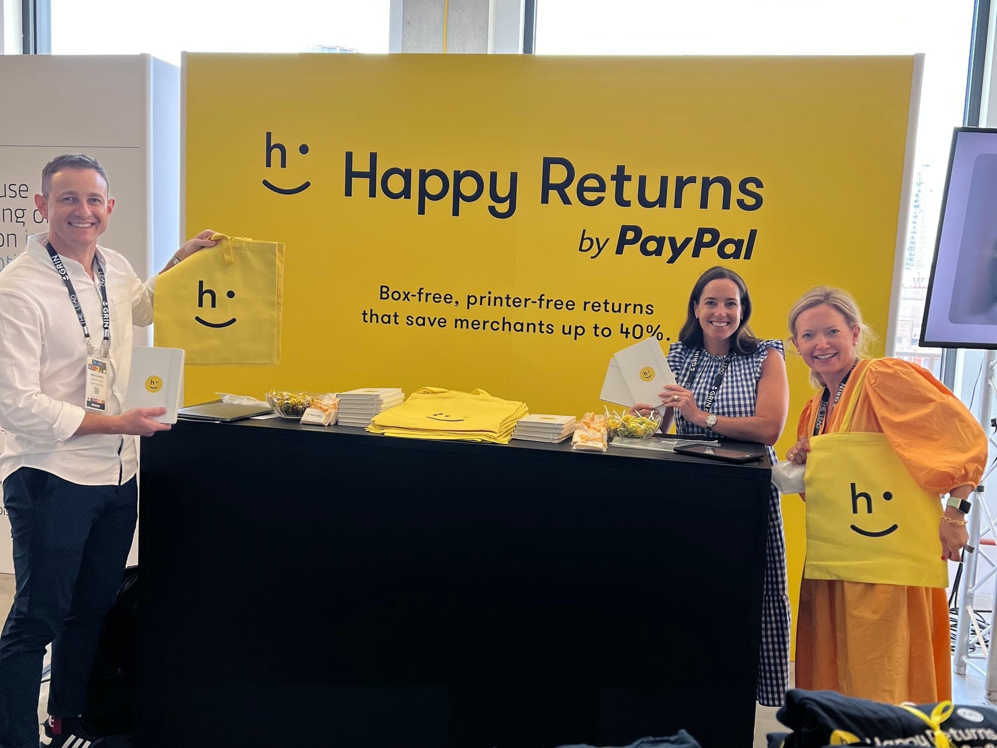 Three people at a yellow Happy Returns by PayPal trade show booth, holding branded tote bags and notebooks.