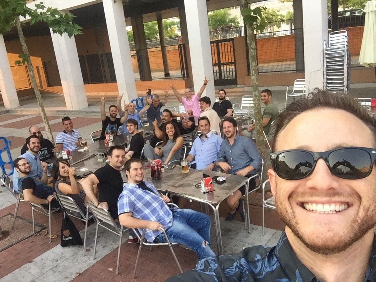 A selfie in front of the team gathered around outdoor café tables, several raising their hands.