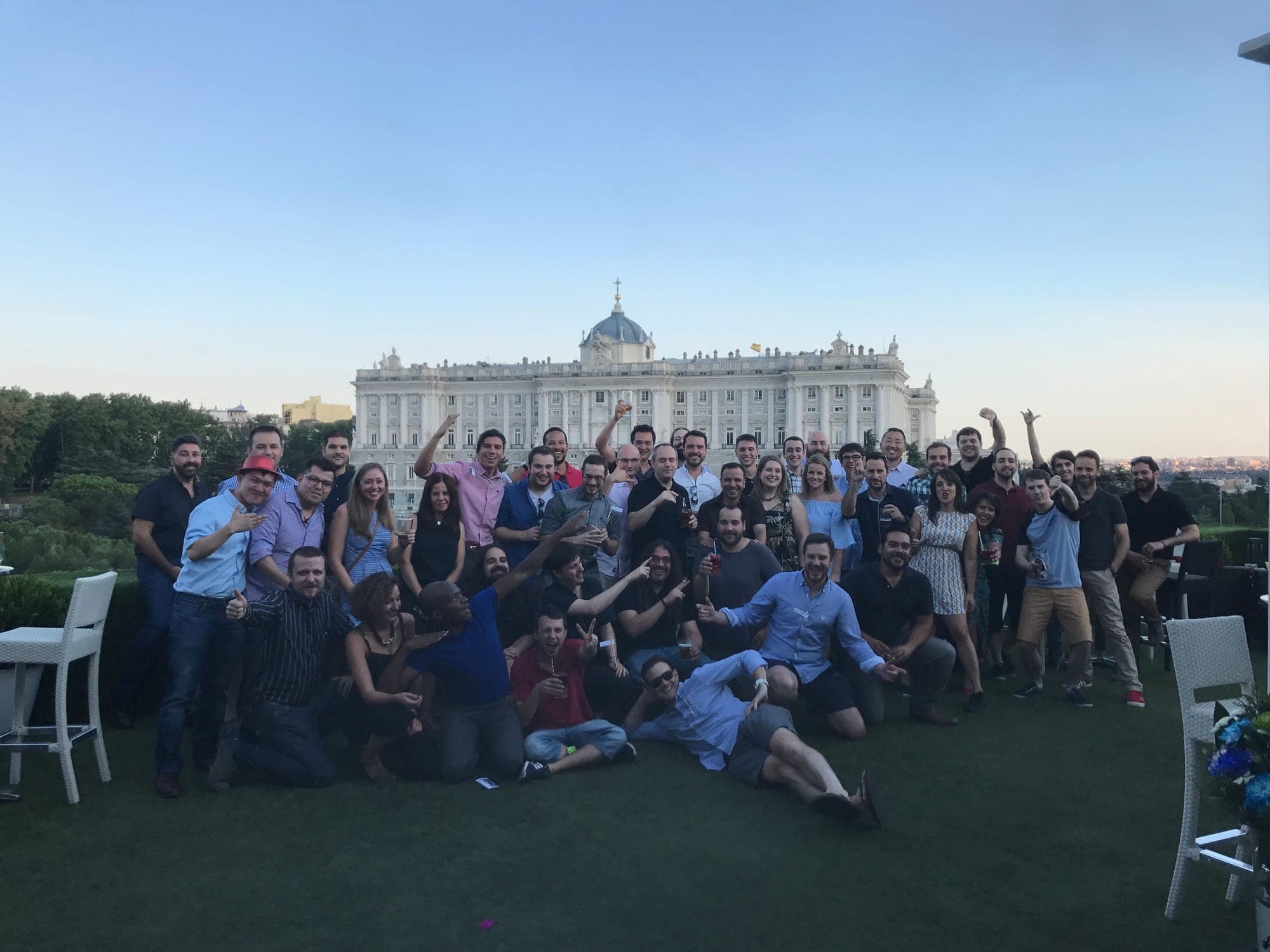 About fifty people posing on a rooftop terrace with the Royal Palace of Madrid behind them.