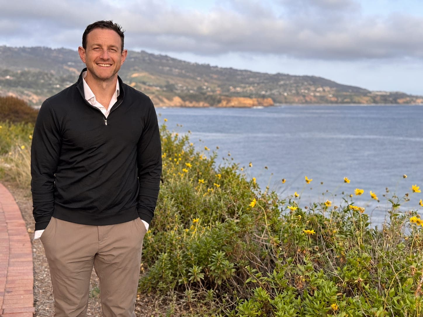 Mike Schwartz smiling on a coastal path, with the ocean, hills and yellow wildflowers behind him.
