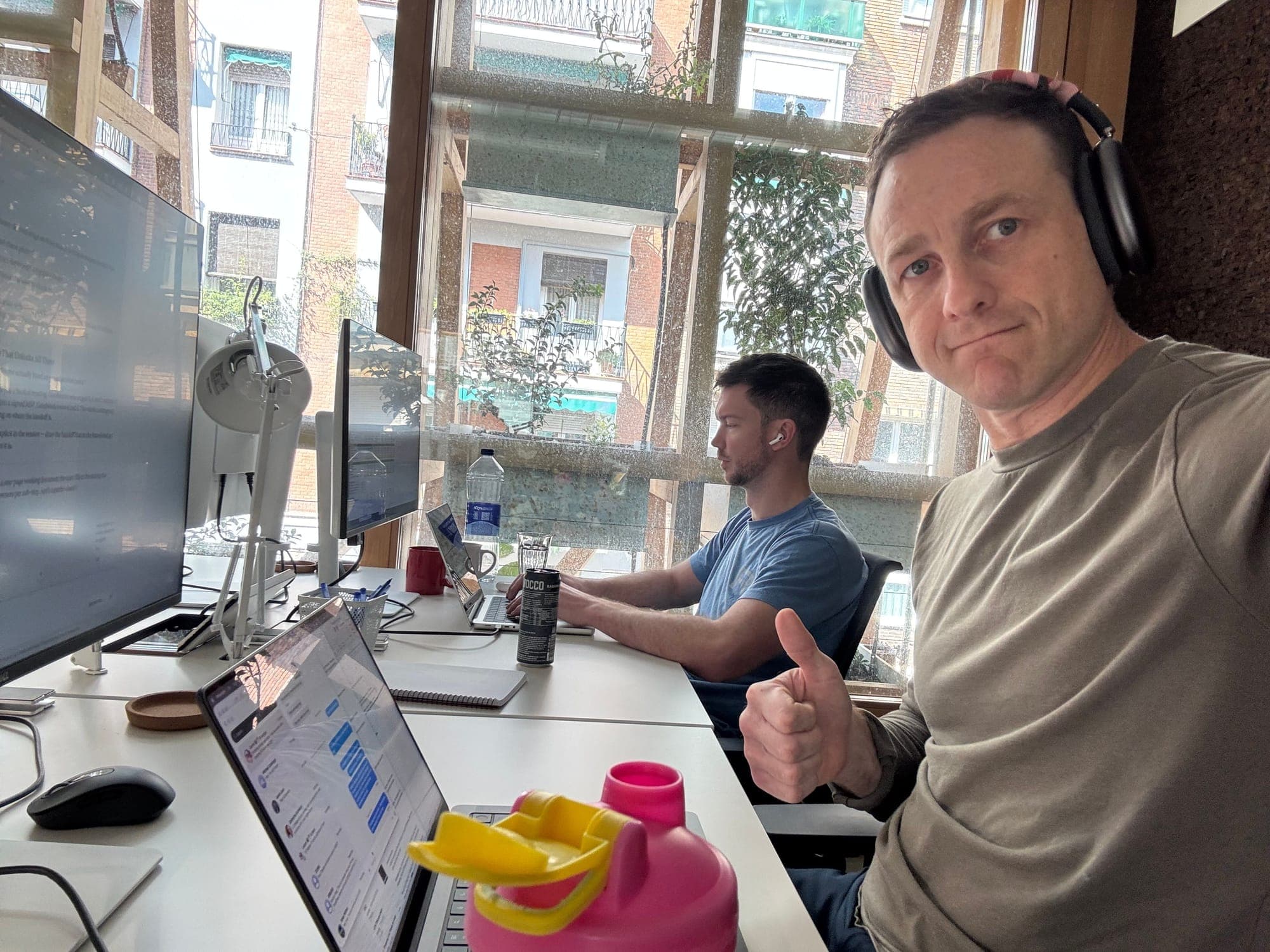 A selfie at a desk with two monitors and headphones, a colleague working at the next desk.